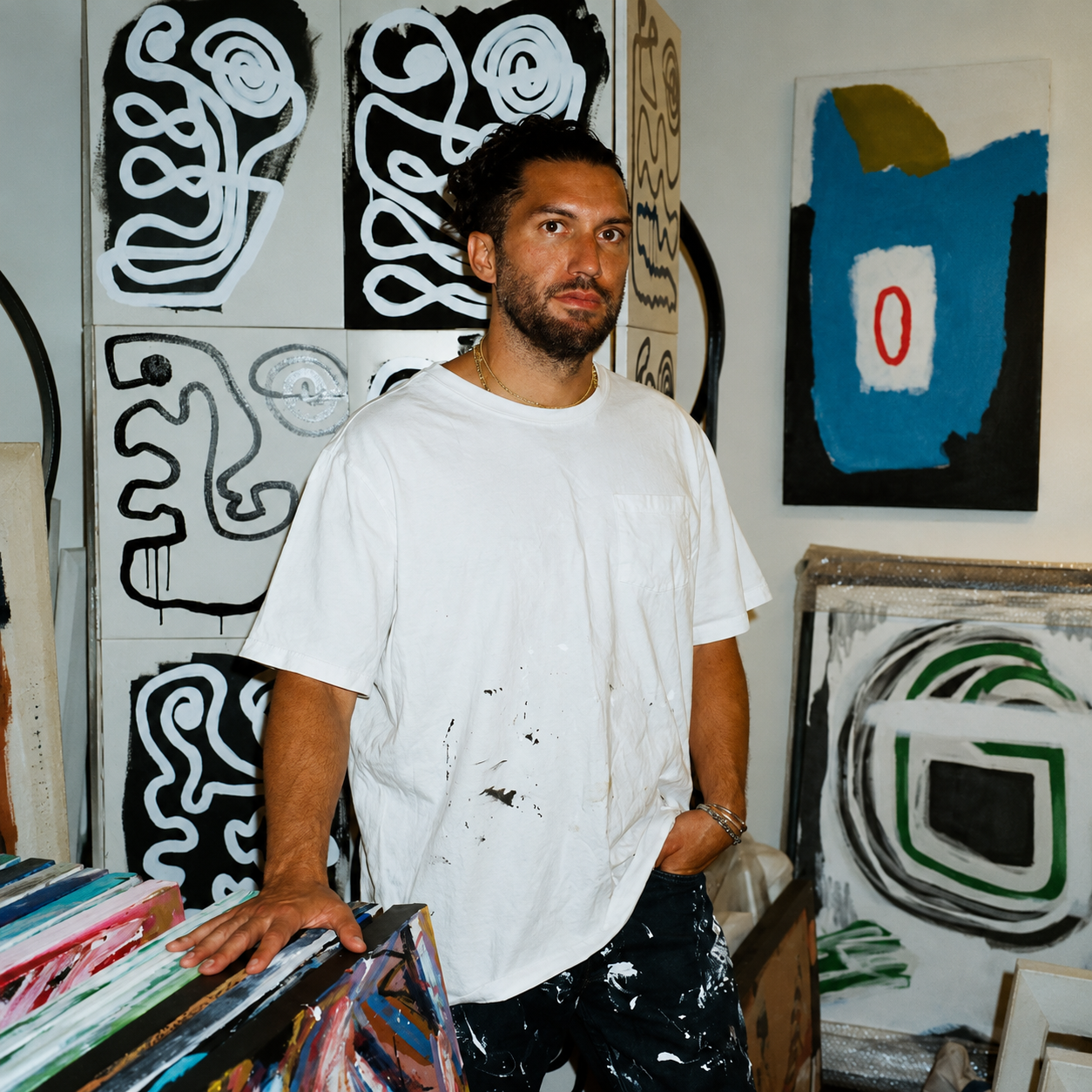 Los Angeles-based artist Dominic J. Lopez stands in his studio wearing a paint-splattered white T-shirt, surrounded by colorful abstract paintings featuring bold, expressive lines and shapes.